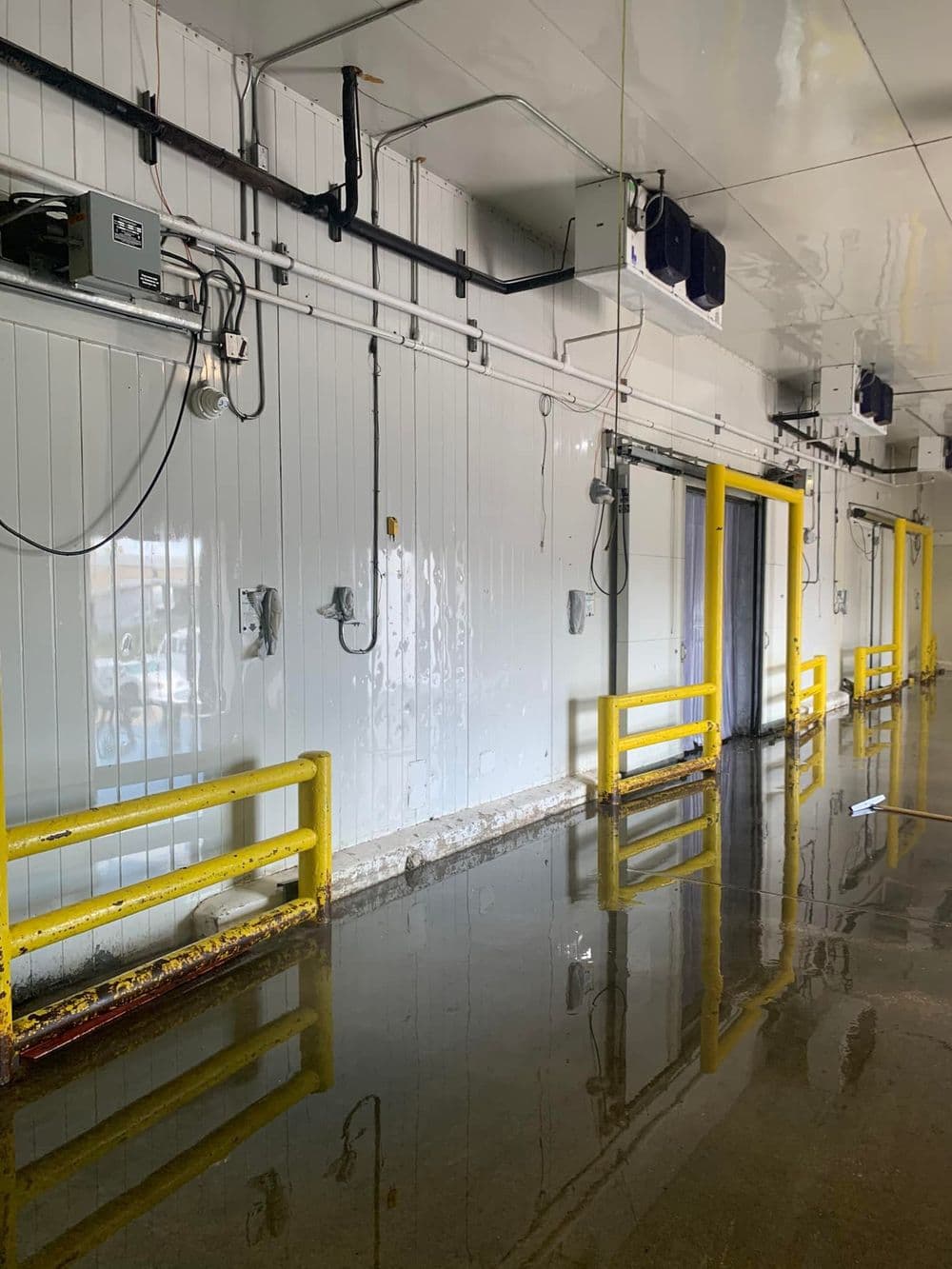 Industrial hallway with water on the floor, featuring yellow safety barriers and wall-mounted equipment.